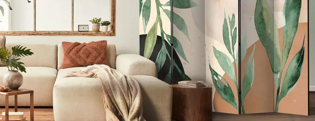 A modern living room with a sofa featuring a decorative cushion and blanket. Next to the sofa stands a decorative room divider adorned with a watercolour pattern of green leaves against a background of beige and black splashes. A wooden coffee table can be seen in the background.
