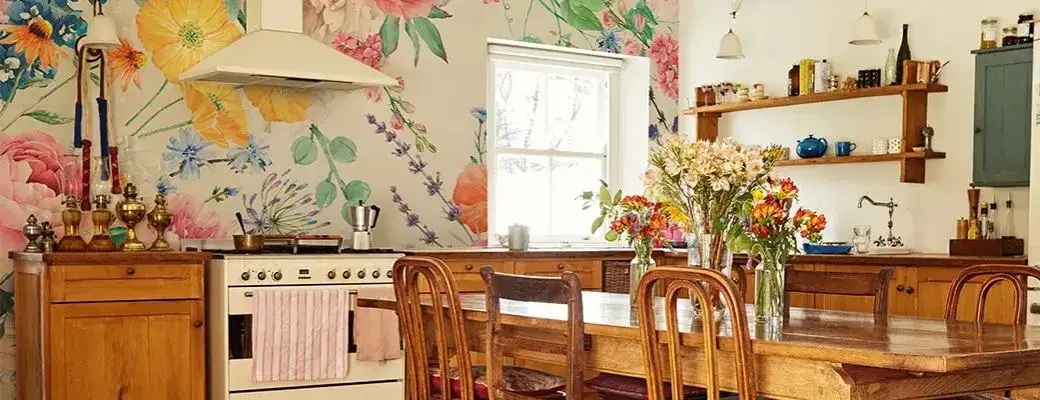 A rustic style kitchen with a large wooden table and chairs. The main feature is the wall above the worktop, covered with a colourful floral wall mural. Vases of fresh flowers stand on the table.