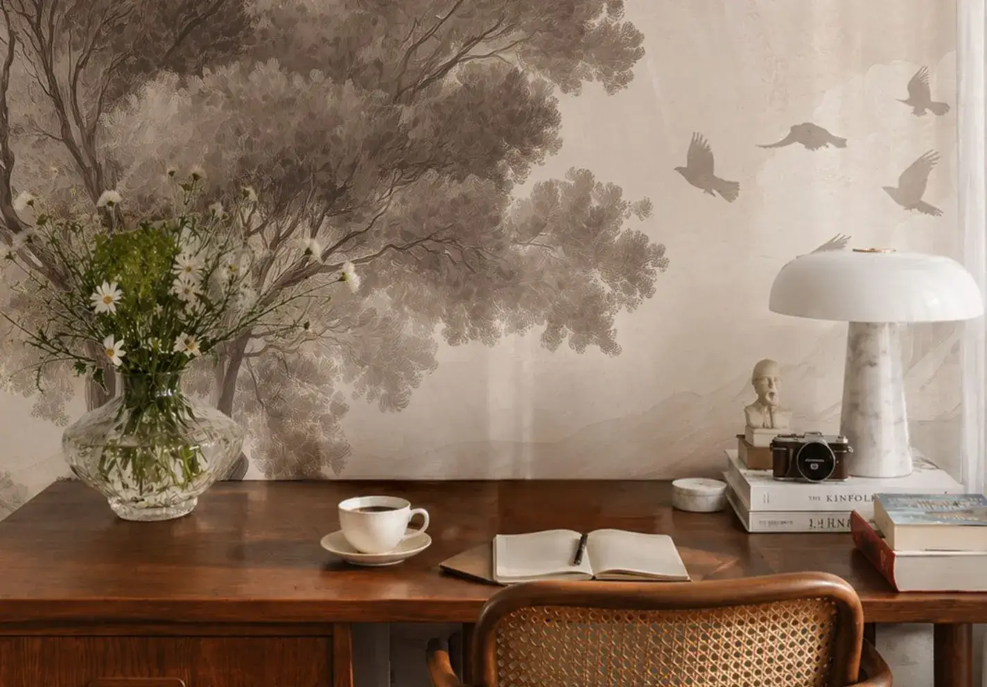 A retro style wooden desk set against a wall featuring a wall mural depicting a large, spreading tree and a flock of birds in flight, rendered in muted shades of sepia and beige. On the desk top lies a notebook, a cup of coffee and a vase of wildflowers.