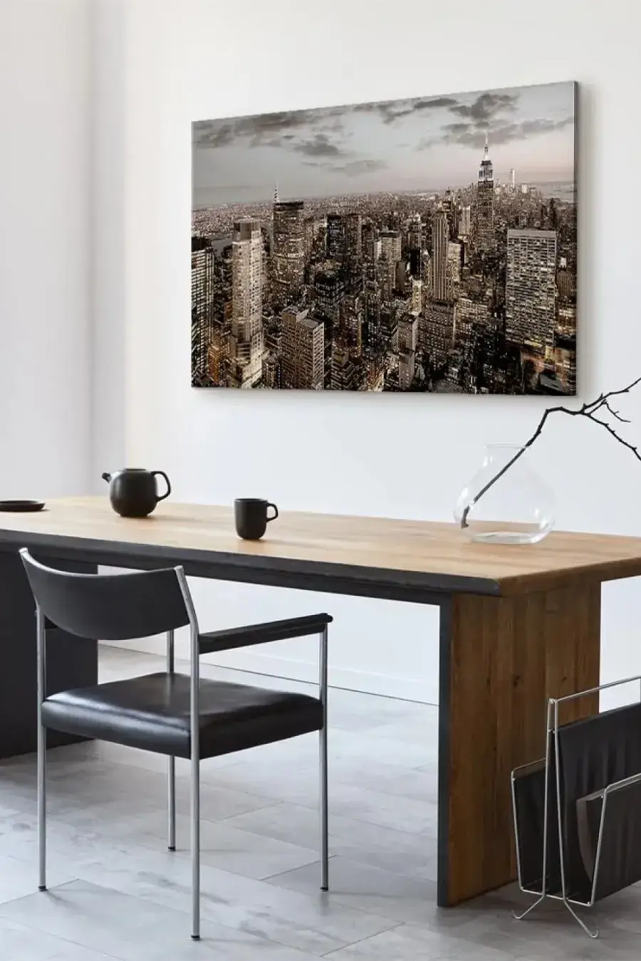 A modern study decorated in a minimalist style. On the wall hangs a large painting depicting New York lit up at night from a bird’s-eye view, with the Empire State Building clearly visible. Below stands a solid desk with a wooden top.