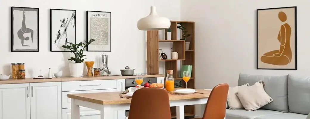 A bright living room with a kitchenette, a dining table and a grey sofa, decorated with modern paintings in black frames on the wall.
